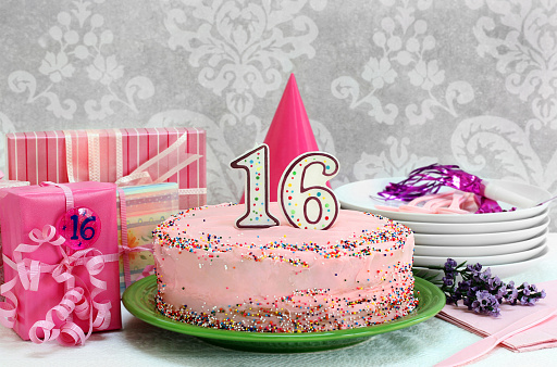 Pink birthday cake with colorful sprinkles and a number 16 candle. Cake is surrounded in gifts, serving plates and flowers.