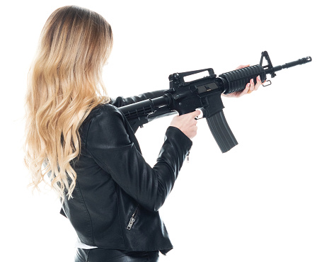 Rear view of who is beautiful with blond hair generation z young women standing in front of white background wearing shirt who is in concentration who is shooting - crime and holding weapon and using machine gun