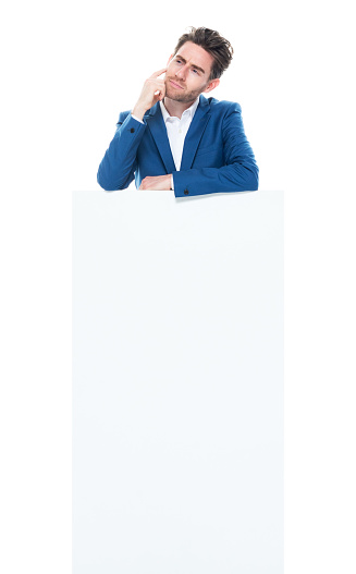 Full length of aged 30-39 years old with brown hair caucasian young male business person standing in front of white background wearing shirt who is asking who is showing with hand and holding placard with copy space