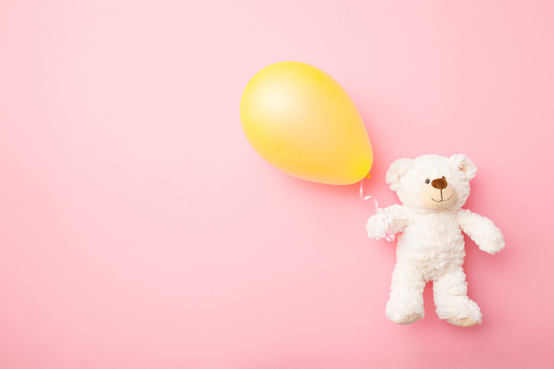 Smiling white teddy bear holding one yellow balloon. Light pink background. Pastel color. Empty place for inspirational, emotional, sentimental text, lovely quote or sayings for good mood. Top view.