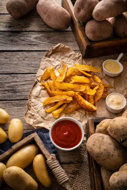 French fries potatoes homemade on a rustic wooden board table with raw potatoes around and sauces ketchup mustard and mayonnaise