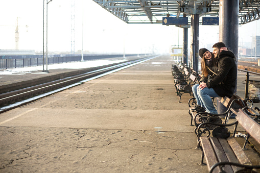 Loving couple sitting on wooden bench at train station. Couple sitting waiting for the train at platform.