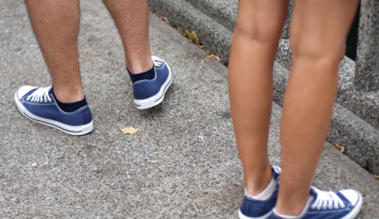 A man and woman wearing matching blue canvas sneakers, standing on a concrete sidewalk. Focus on the back of his left shoe, her legs and feet are deliberately out of focus.