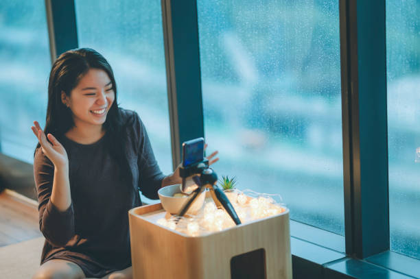 アジアの中国のティーンエイジャーの女の子が彼女の部屋で彼女の部屋で、彼女のスマートフォンを使って、笑顔でリンゴを食べてブログを書く - ビデオブログ ストックフォトと画像