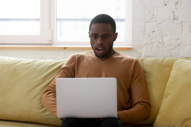 African ethnicity guy gawp at laptop screen feels shocked African guy seated on sofa looks at laptop screen feels shocked by got incredible business opportunity. Stunned American man makes big eyes open mouth gawp at pc monitor, unbelievable or awful news gawp stock pictures, royalty-free photos & images