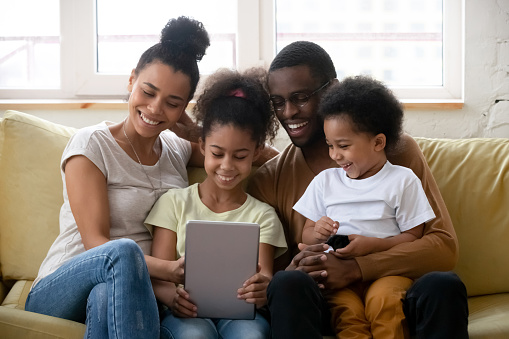 Beautiful full African family spend free time using tablet device watching cartoon educational app for kids enjoy on-line services making videocall chatting with relatives sit on couch in living room