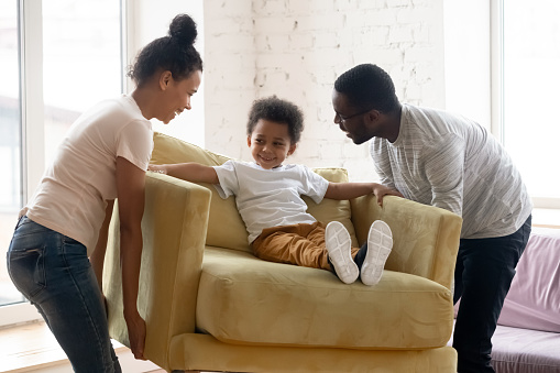 African couple carrying armchair where sit adorable small toddler son. Happy family placing delivered furniture bought in modern store, furniture shop advertisement, relocation day at new home concept