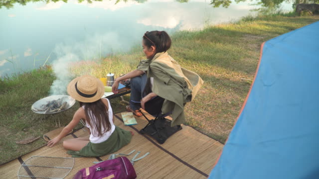 rear view of Asian mother and mixed-race daughter travel camping together in a forest, Happy and relaxed woman and teenage girl cooking BBQ is in front of a tent. Concept of adventure family at the weekend.