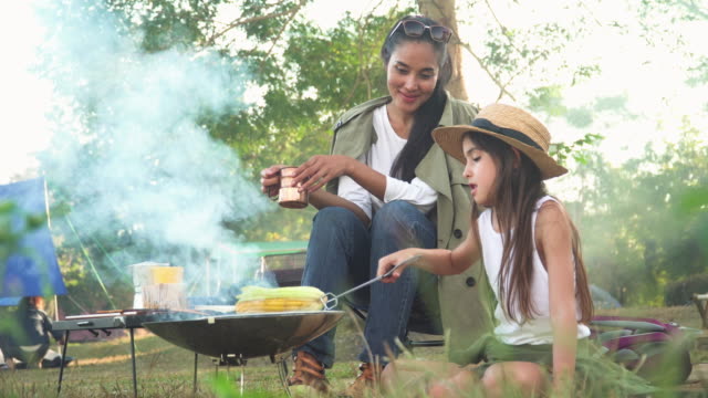 front view of Asian mother and mixed-race daughter travel camping together in a forest, Happy and relaxed woman and teenage girl cooking BBQ is in front of a tent. Concept of adventure family at the weekend.