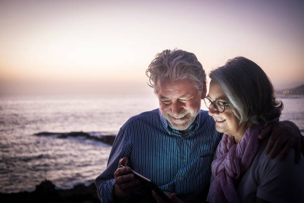 istock Happy retired couple having fun sitting on the cliffs, dusk light looking at the same tablet. Happy retirement concept. Sea in the background 1247055338