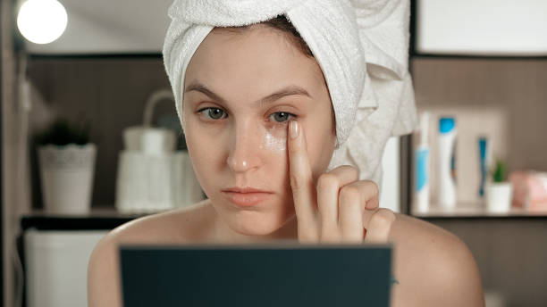 Eye cream. Young attractive woman applies cream to skin under her left eye, girl is in bathroom and looks in mirror. Hygiene, beautician, makeup, face care concept. Close-up Eye cream. Young attractive woman applies cream to skin under her left eye, girl is in bathroom and looks in mirror. Hygiene, beautician, makeup, face care concept. Close-up view slow motion face stock pictures, royalty-free photos & images