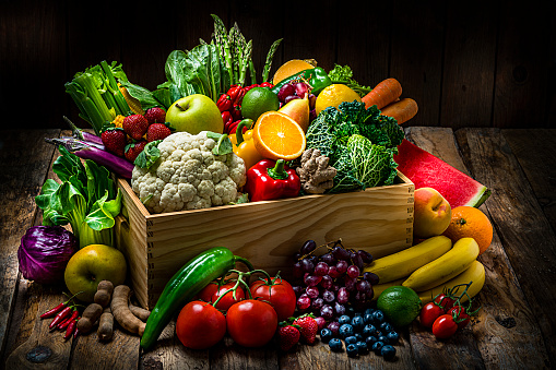 Healthy food: large selection of fresh organic multicolored vegetables in a crate shot on dark wooden table. Fruits and vegetables included in the composition are watermelon, oranges,lime, lemons, banana,grape, strawberries, apples, pears, blueberries, tamarind, kale, tomatoes, squash, asparagus, celery, eggplant, carrots, lettuce, edible mushrooms, bell peppers, cauliflower, ginger, corn, bok choy, raw potatoes, chili peppers among others. High resolution studio digital capture taken with Sony A7rII and Sony FE 90mm f2.8 macro G OSS lens