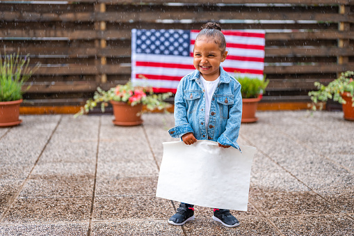 Two years old cute little african-american girl, wearing white t-shirt and jeans jacket, standing in the rain and holding a white poster. She is smiling and looking away. American flag on the wooden fence and flower pots in the blurred background.