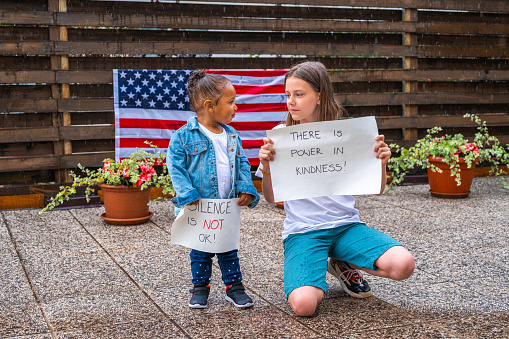 Young caucasian girl and a little african-american girl standing outdoors in the rain. Bigger caucasian girl on the right is wearing white t-shirt, short blue pants and sneakers and she is holding a white poster with handwritten message on it: THERE IS POWER IN KINDNESS! And a small girl on the left, wearing a white t-shirt, jeans jacket, blue leggings and sneakers is holding a white poster with handwritten message: VIOLENCE IS NOT OK! They are looking at each other. There is an american flag on the wooden fence and some flower pots in the blurred background.