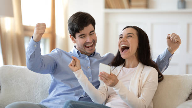 Overjoyed couple triumph winning lottery on cell Excited young couple triumph winning online lottery on smartphone gadget, overjoyed man and woman relax at home feel euphoric get read unexpected great news or sale offer on internet on cellphone unusual couple stock pictures, royalty-free photos & images