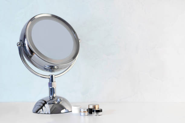 хромированное стальное круглое увеличительное зеркало на ручке, настроенное на белом столе с некоторыми банками со сливками и косметикой. - table optical instrument metal steel стоковые фото и изображения