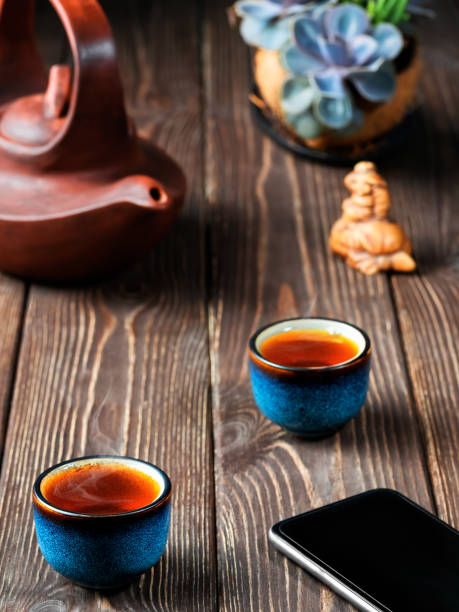 istock Two bowls with red tea, clay teapot on the table, tea ceremony. Smartphone on the table next to the bowls. Freshly brewed black tea, steam rises above the cups, dark wooden background, close-up 1247256063