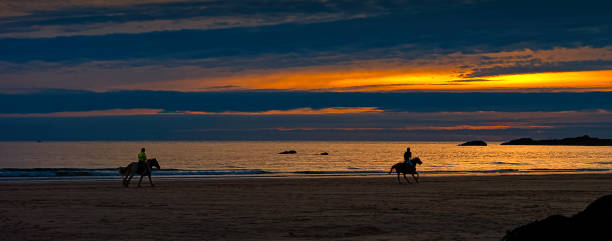Jaco, Costa Rica- February 18, 2023: people enjoying the sunset on the beach while a cowboy leads two riders on horses