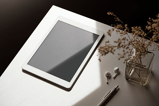 White blank screen digital tablet mockup, template at the corner of the table with natural light and shadow. Flowers in the vase, earphones and pen on the table.
