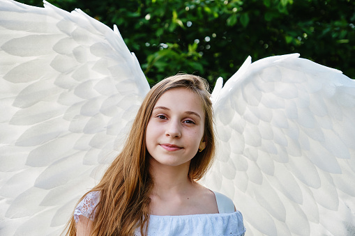 cute white teenage girl with angel wings and white dress posing outdoors in park.