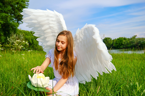 young beautiful white girl with angel wings plays with waterlily sitting on a meadow in the grass near the riverside. Sunny summer day