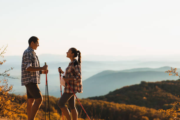 young pregnant woman with boyfriend nordic walking outdoors with trekking poles.  active and healthy lifestyle in maternity time. beautiful autumn mountain view. - couple human pregnancy sunset walking imagens e fotografias de stock