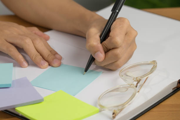 istock A close up of a white woman's hand Writing note Attached to a white notebook and placed on a wooden desk and with glasses placed 1247447197