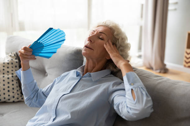 istock Overheated elderly female leaned on couch refreshing herself with fan 1247568825