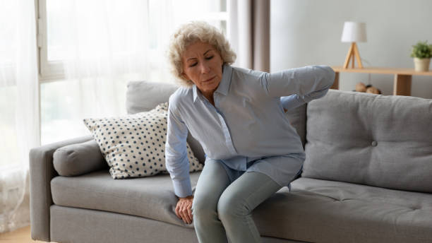old woman gets up off the sofa feels repeated low backpain - low back imagens e fotografias de stock