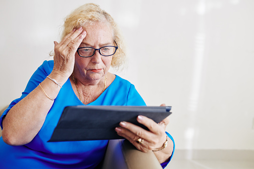 Confused senior woman in glasses reading disturbing news on tablet computer