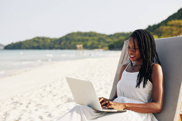 Woman resting in chaise-lounge Portrait of pretty smiling young Black woman resting on chaise-lounge on sandy beach and working on laptop digital nomad stock pictures, royalty-free photos & images
