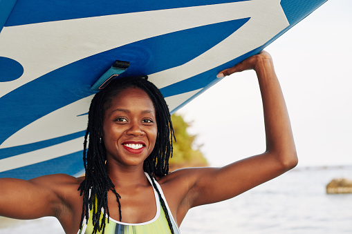 Portrait of happy wet young Black woman carrying surfboard over her head
