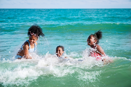 Happy three African American kids are swimming in the sea playfully together. With waves and splashes of water. Happy friendship. Happy vacation holiday. Relaxation in vacation in the summer concept.