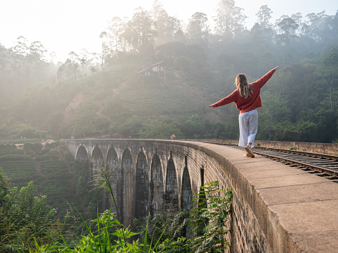 Playful young woman walking on the edge of ancient viaduct bridge at sunrise and balancing with arms outstretched by her side. Girl travel fun Sri Lanka