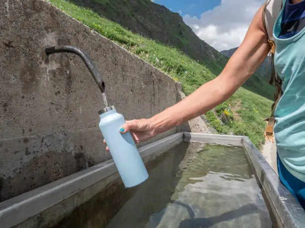 Unrecognisable woman on mountain trail filling up water bottle and drinking the fresh water from the valley at the source; Girl hiking in Summer refreshing