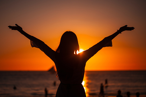 Silhouette of woman raising arms to golden sunset over sea. Freedom, success and hope concept. Girl relaxing and enjoying peace and serenity on beautiful nature, vacation concept.