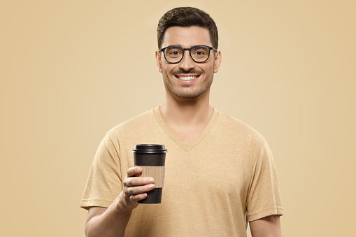 Young handsome man wearing beige t-shirt and eyeglasses, holding takeaway coffee cup in one hand, satisfied with free time, isolated on brown background