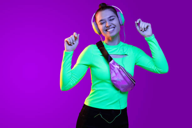 joven chica feliz usando top verde neón, bolso de cinturón violeta y bailando música que está escuchando a través de auriculares, aislado sobre fondo púrpura - purple belt fotografías e imágenes de stock