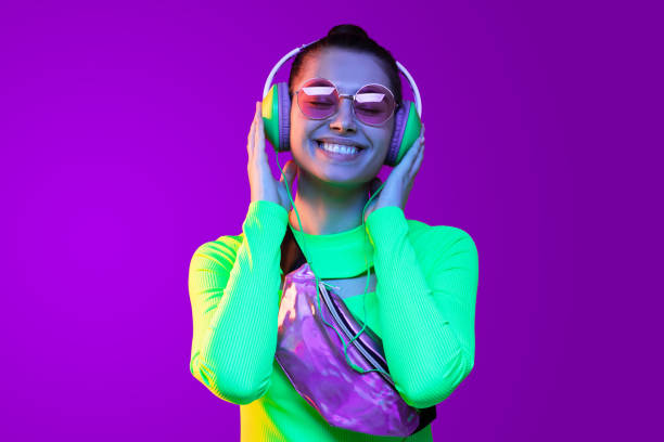 retrato de cerca de la joven feliz en la parte superior verde neón, con bolsa de cinturón, gafas y auriculares, escuchando la canción favorita con la sonrisa, aislado en el fondo púrpura - purple belt fotografías e imágenes de stock