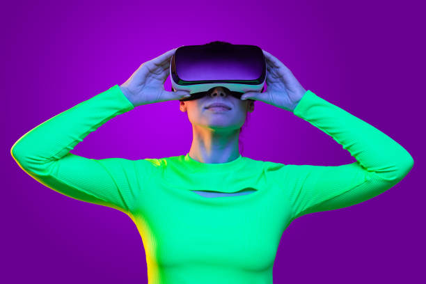 istock Young female in neon green top wearing virtual reality headset and holding it with both hands, watching new gaming world, isolated on purple background 1247694607
