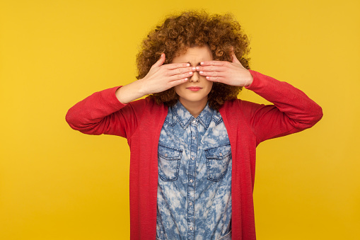 Don't want to look. Portrait of scared woman with curly hair in casual outfit covering eyes, feeling shy and afraid to watch, ignoring troubles. indoor studio shot isolated on yellow background