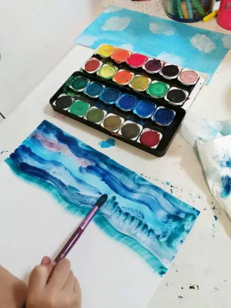 Watercolor palette and kid's hand holds a brush to paint on white paper. Concept keep the sea clean from plastic, safe the planet. kid's education