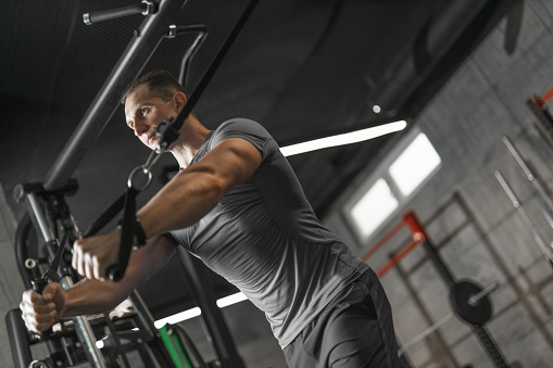 Handsome sporty man works out pushing up exercises on cable crossover and flexing muscles in health club