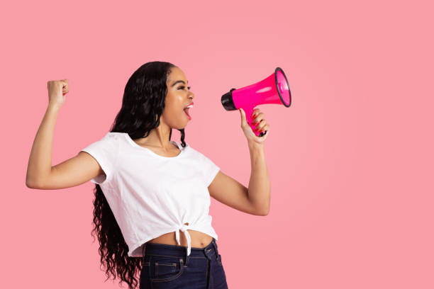 verticale d’une jeune femme étant bruyante et entendue en criant par le mégaphone avec le poing vers le haut et la bouche ouverte - screaming shouting women using voice photos et images de collection