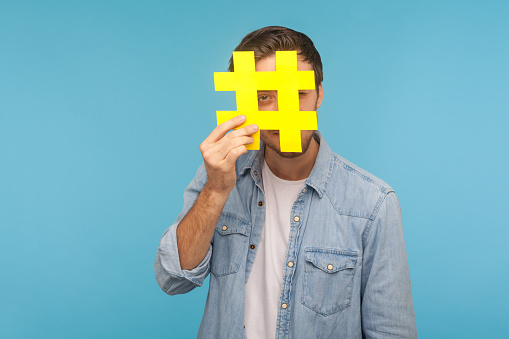 Portrait of man in denim shirt looking through big yellow hashtag symbol, standing isolated on blue background with copy space for tagged message, popular internet idea, viral content. studio shot