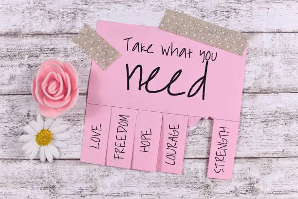 Pink tear-off stub note with text 'Take what you need' and words 'Love, Freedom, Hope, Courage' and 'Strength' and one stub missing with cute flowers on light wooden background