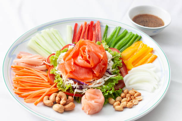 Chinese new year auspicious food (Yu Sheng) eating on celebrations ingredients with peanut, salmon, nut, red ginger, sliced carrot, sliced chili, sliced Cucumber, sliced tomato on white dish and served with sauce. comida china de año nuevo auspicioso (yu sheng) comiendo en celebraciones ingredientes con cacahuete, salmón, nuez, jengibre rojo, zanahoria en rodajas, chile en rodajas, pepino en rodajas, tomate en rodajas en plato blanco y servido con salsa.
