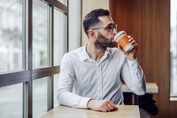 istock Young handsome bearded elegant man sitting in cafeteria and drinking fresh morning coffee from disposable cup. 1247754604