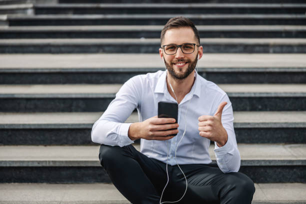 若い魅力的なひげを生やしたエレガントなビジネスマンは、彼の会社の前の階段に座って、スマートフォンを使用し、親指を見せています。 - high society audio ストックフォトと画像