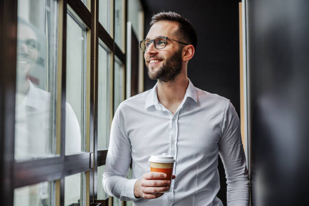 istock Young attractive bearded businessman standing neat window on coffee break, looking trough it and holding fresh morning coffee. 1247754943